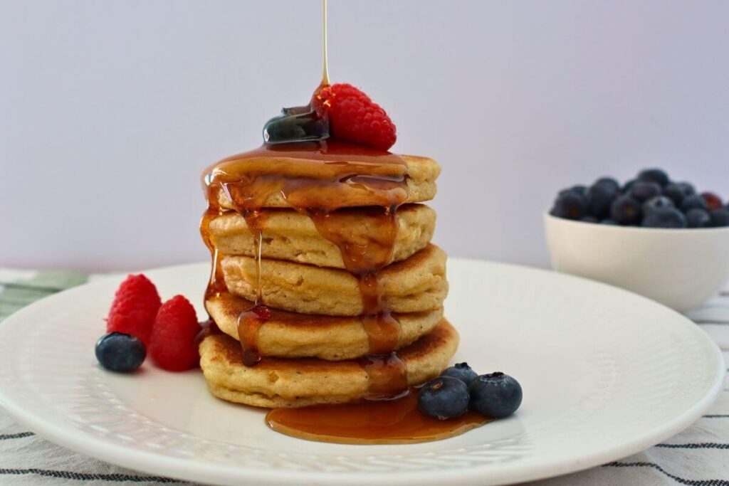 Stack of 5 fluffy almond flour griddle pancakes on a plate with berries and maple syrup drizzled over top and a bowl of blueberries in the background