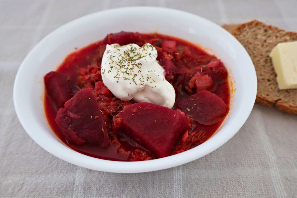 from-scratch beef borscht in a bowl with sour cream and herbs on top and a slice of bread and butter in the back round