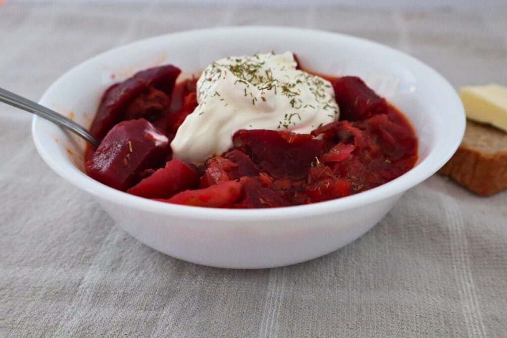 bowl of borscht soup with sour cream and herbs on top and a spoon in the soup with bread and butter in the back round