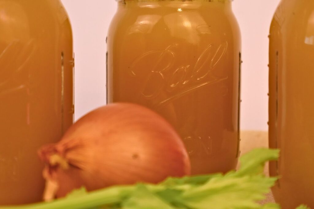 Closeup of 3 mason jars full of chicken bone broth with celery and an onion.