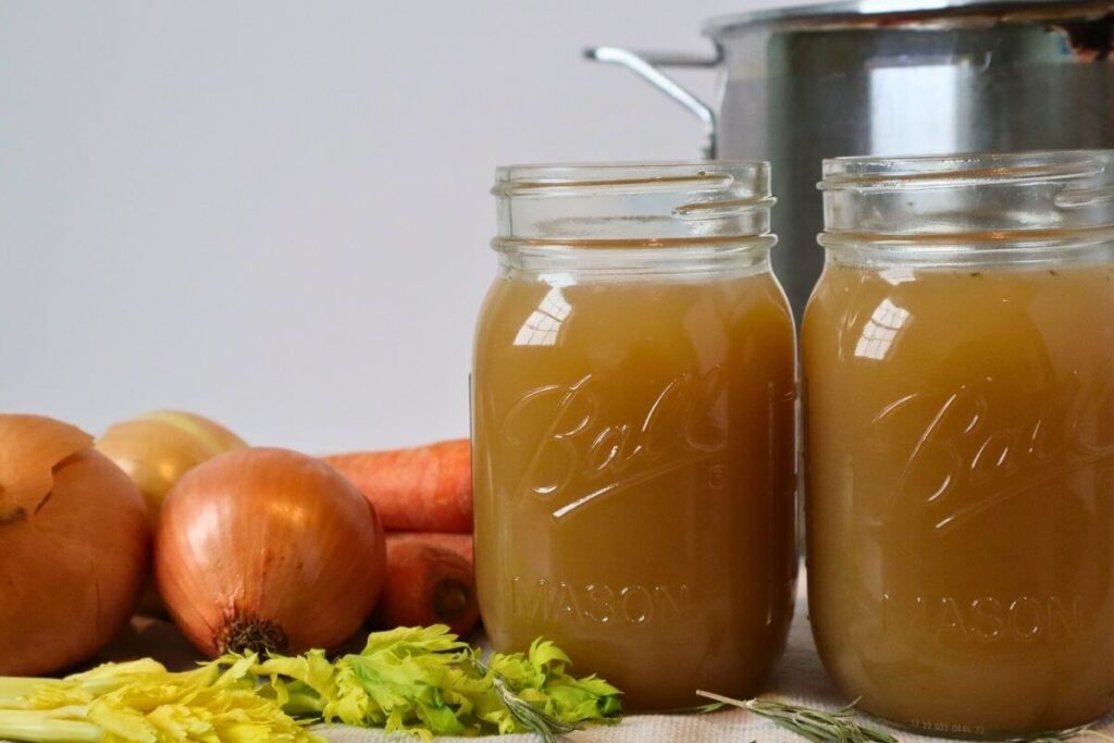 2 jars of finished beef bone broth on the stovetop with vegetables and a stockpot