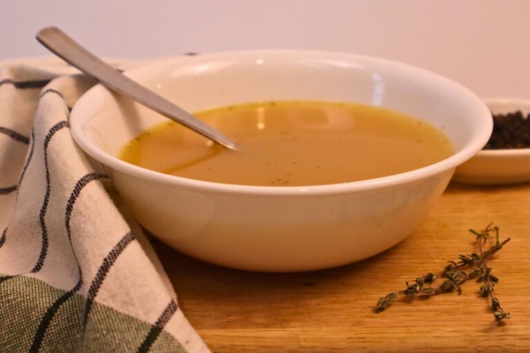 Chicken bone broth in a white bowl with a spoon and herbs.