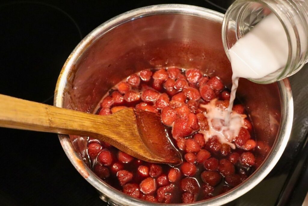 Adding arrowroot powder to a kettle of homemade strawberry sauce to thicken it.