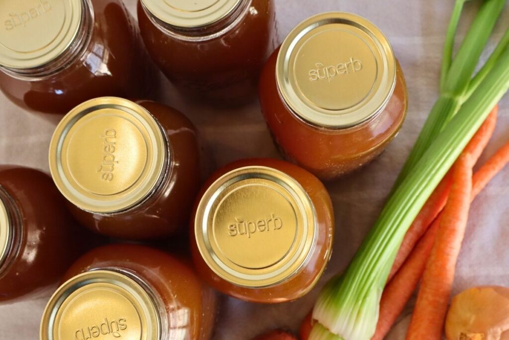 Overhead view of 7 jars of golden bone broth with onion, carrot, and celery stalks.