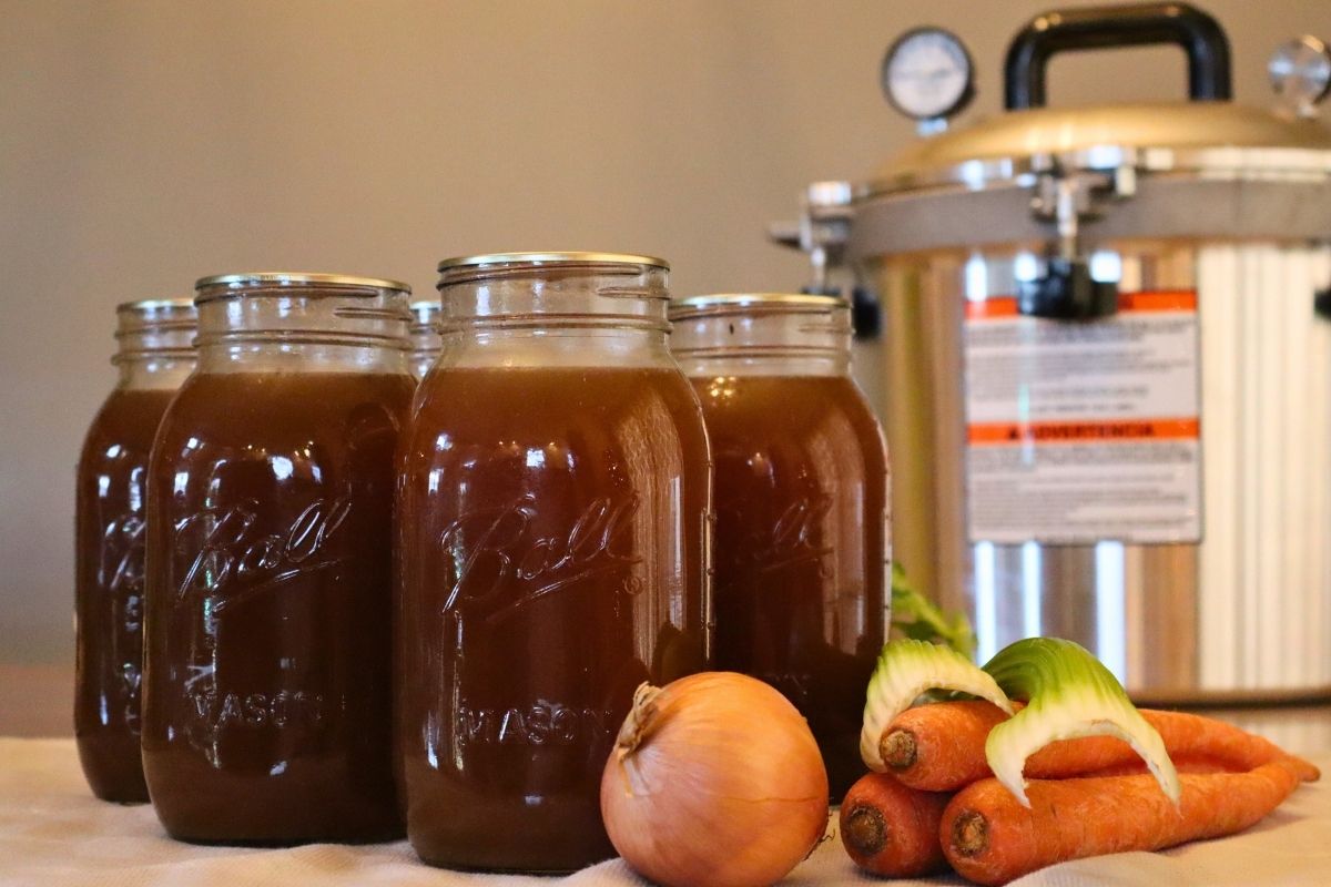 Closeup of 5 jars of nutritious bone broth with onion, carrots, and celery with a pressure canner in the background demonstrating how to pressure can bone broth.