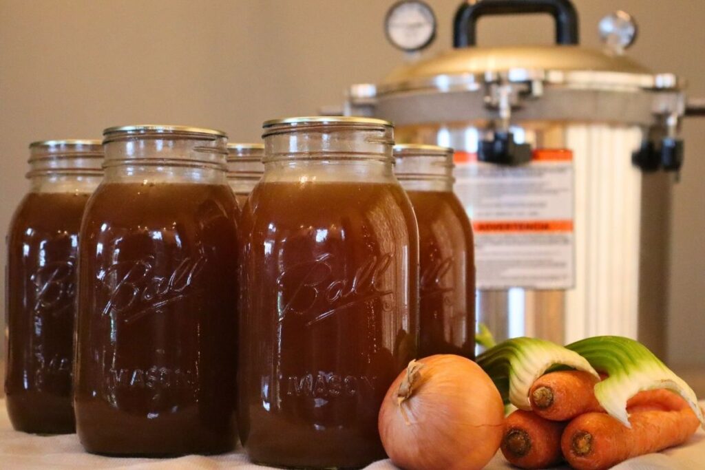 5 jars of nutritious bone broth with onion, carrots, and celery with a pressure canner in the background demonstrating how to pressure can bone broth.