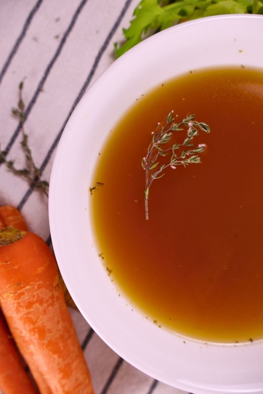Bowl of rich chicken bone broth with a sprig of thyme on top, surrounded by carrots and celery on a tea towel