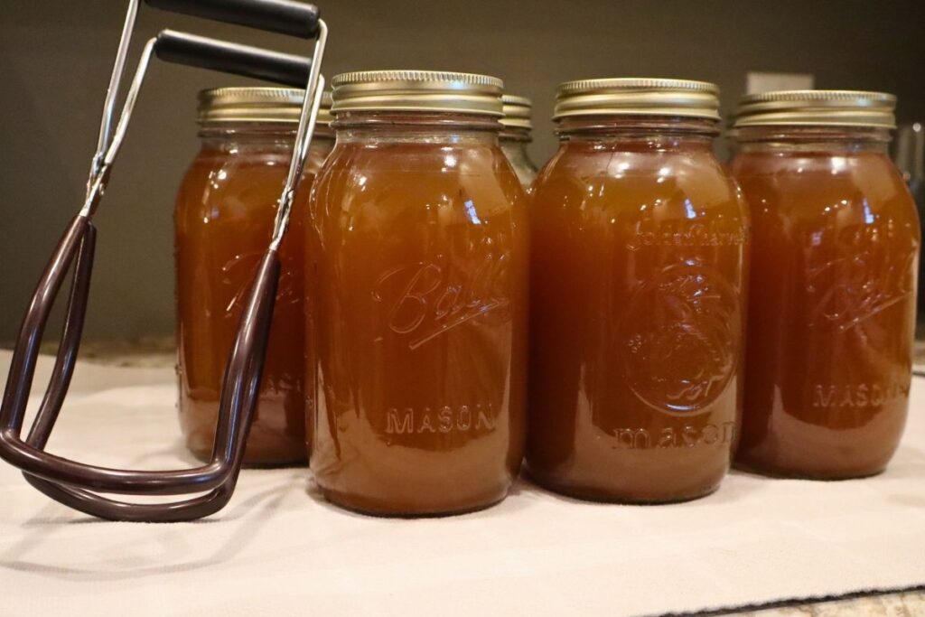 Golden bone broth lined up with a jar lifter cooling after being pressure canned.