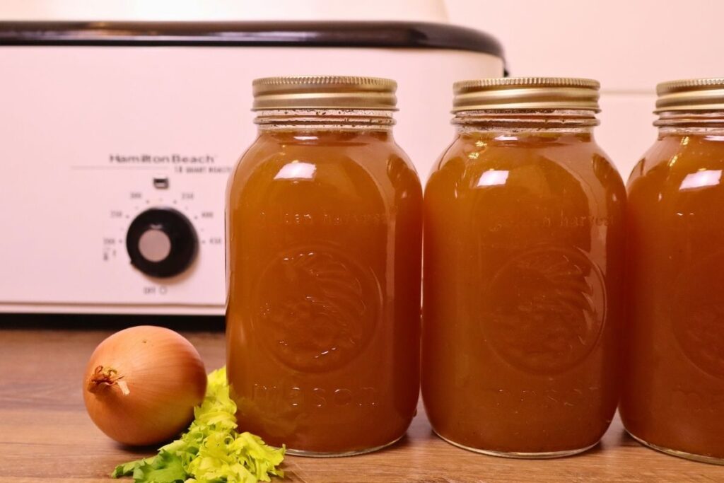 3-quart jars of chicken bone broth with onion and celery and an electric roaster in the background.