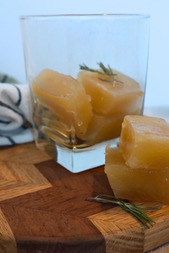 Frozen bone broth cubes in a glass cup sitting on a wooden cutting board sprinkles with dried rosemary. A creative look at how to freeze bone broth for a quick nutrient-dense hot morning drink or easy cooking portions.