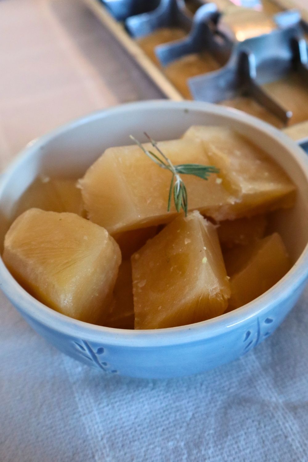 Golden frozen bone broth cubes topped with dried rosemary in a bowl, showing a healthy meal prep hack for how to freeze bone broth for sipping or recipes. A metal ice cube tray sits in the background of this rustic homestead kitchen shot.