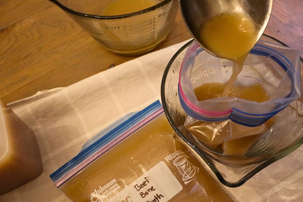 A ladle pouring bone broth into a freezer bag inside a glass bowl with a bag full of bone broth beside it ready to go into the freezer.