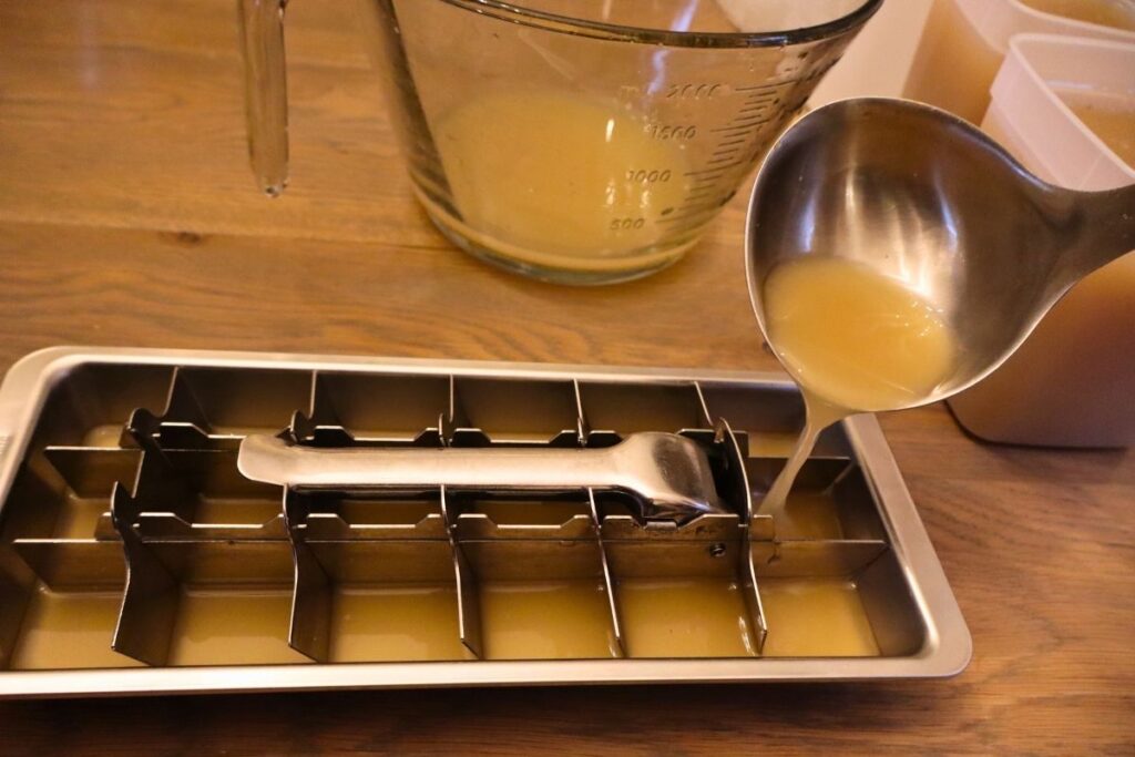 Pouring bone broth into an ice cube tray with containers of bone broth in the background, ready to go into the freezer for storage to use in soups, stews, sauces, and recipes.