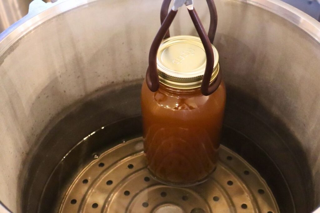 Using a jar lifter to place a jar of bone broth into an empty pressure canner.