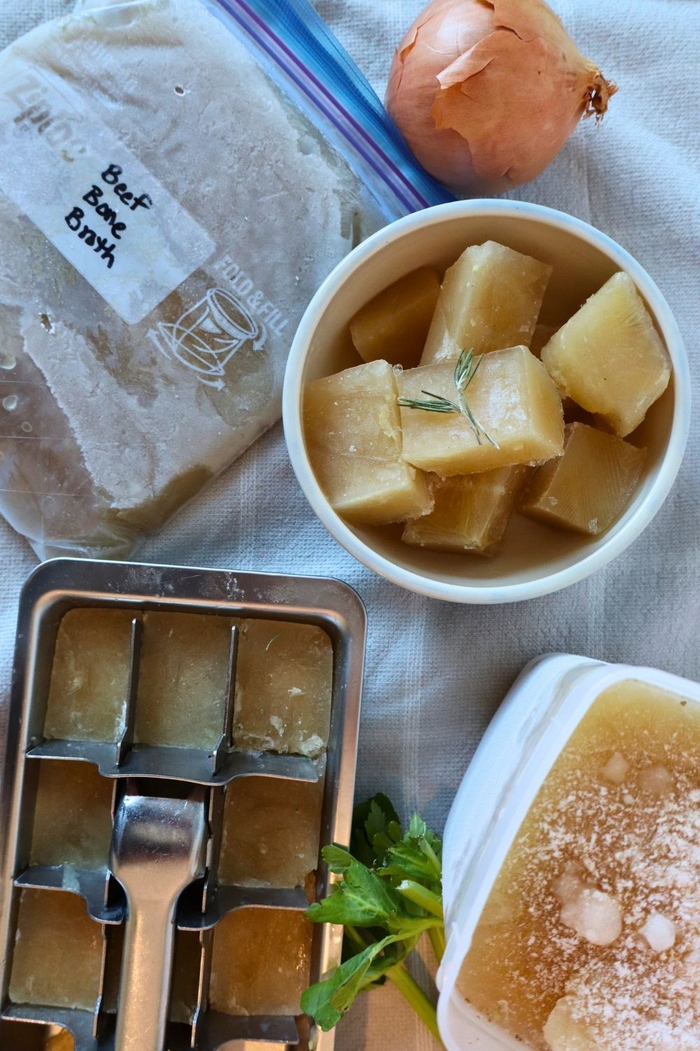 Ice cube tray with frozen bone broth cubes, a bowl of frozen bone broth cubes topped with fresh rosemary, a container of frozen bone broth, and a freezer bag of frozen bone broth with onion and celery, ready for soups, stews, and from-scratch recipes.