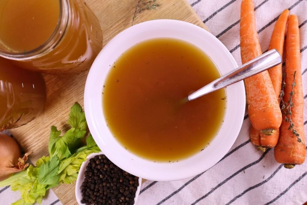 A bowl of homemade bone broth with a spoon, a jar of broth, carrots, and peppercorns — how to make bone broth at home