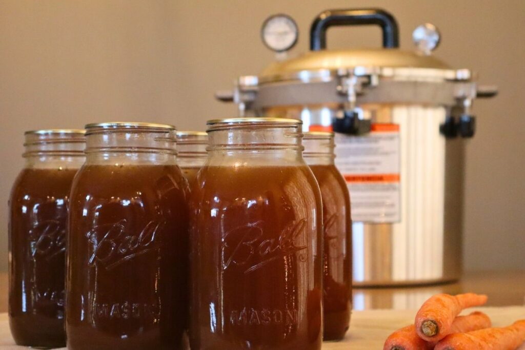 Closeup of 5 jars of nutritious bone broth with carrots, and a pressure canner in the background for a step-by-step guide for how to pressure can bone broth.