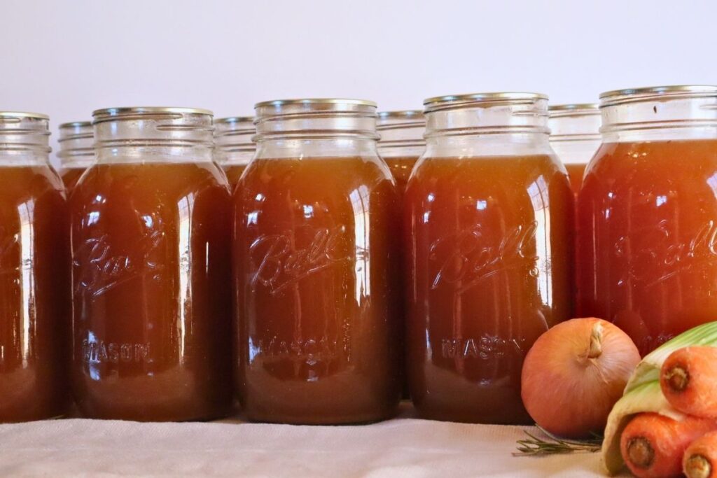 9 jars of rich golden bone broth lined up with onions, celery, carrots, and rosemary.