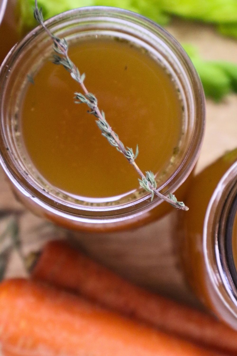 Close-up of homemade chicken bone broth in a mason jar with a sprig of thyme on top. This gelatinous broth was made in an electric roaster for large-batch cooking. Background includes carrots and celery.