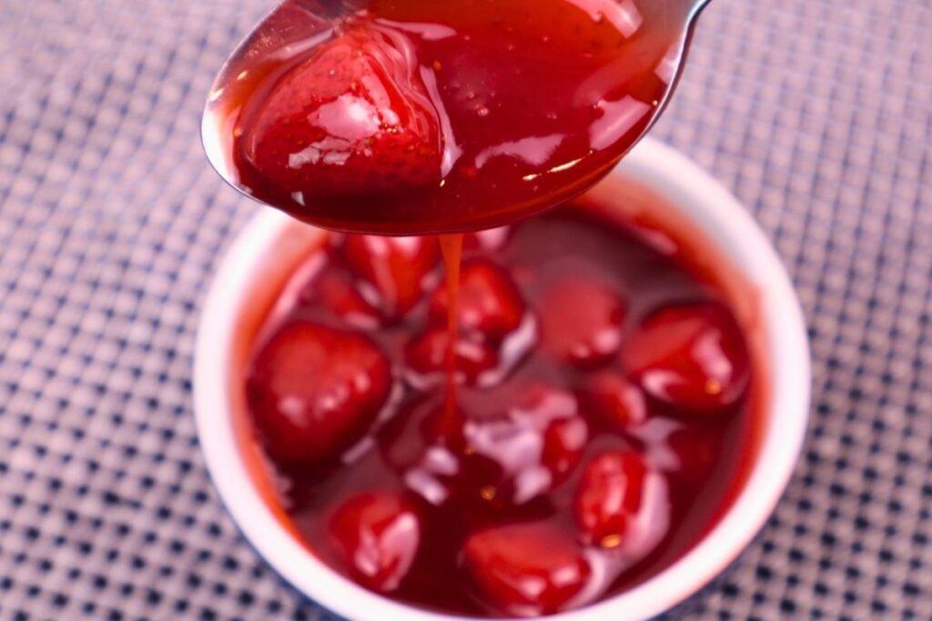 Smooth strawberry sauce dripping from a spoon falling into a white bowl.