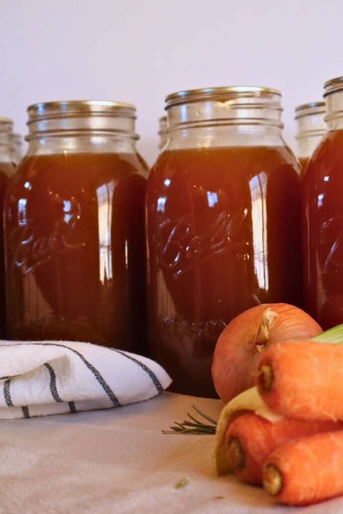 Jars of pressure canned bone broth in the background with carrots, celery, onion, and rosemary.