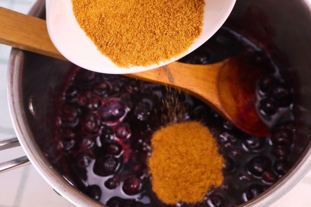 Coconut sugar being poured into a kettle of mostly thawed blueberries.