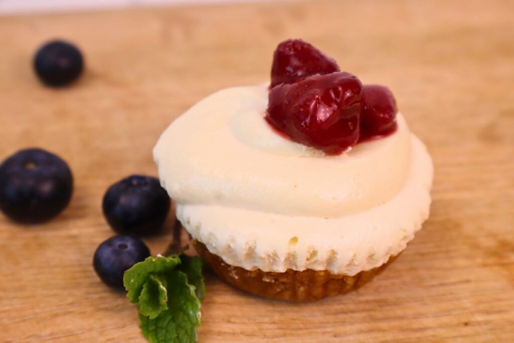 Gluten-Free Mini No-Bake Cheesecakes with Berry Sauce 22 A gluten free mini no bake cheesecake topped with homemade strawberry sauce with fresh blueberries and mint leaves beside it.