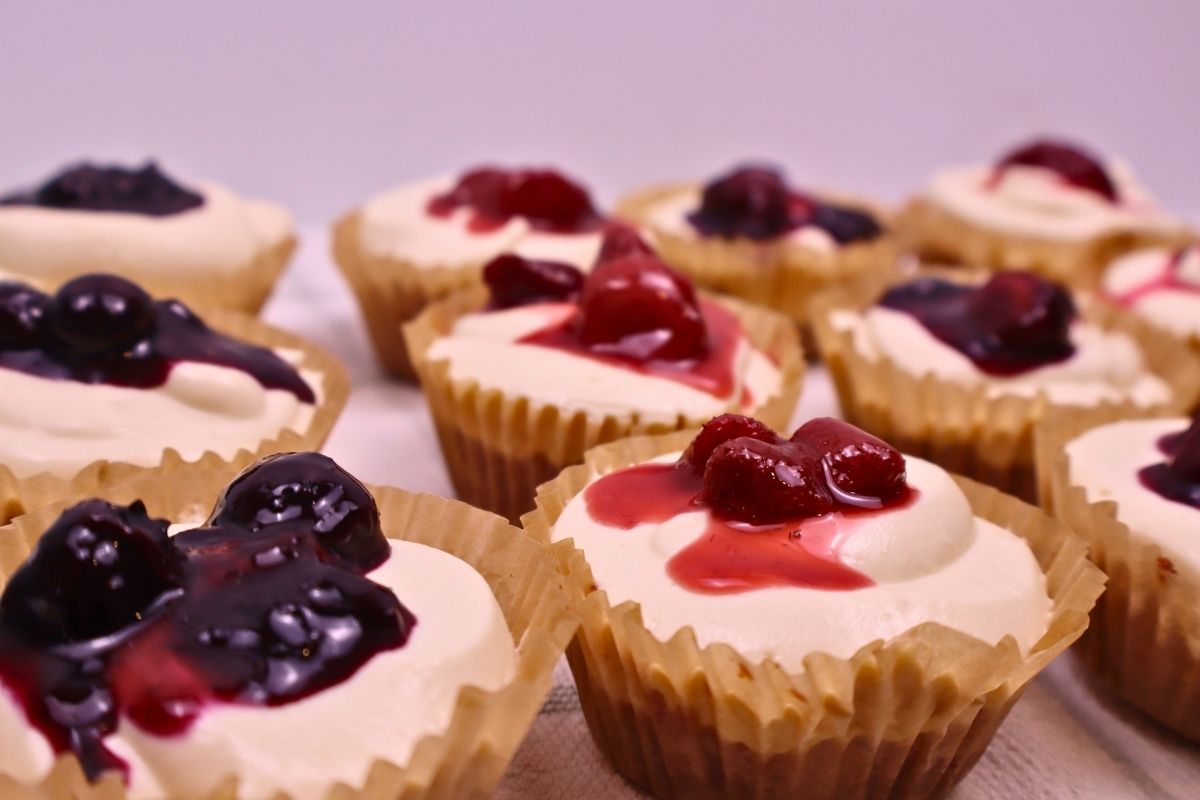 Angled close up view of twelve gluten free mini no bake cheesecakes topped with a variety of homemade mixed berry, strawberry, and blueberry sauces.