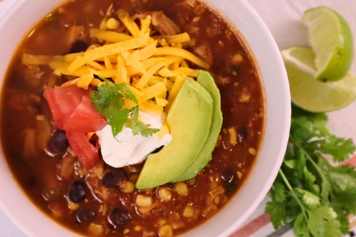 Overhead shot of gluten-free chicken taco soup topped with sour cream, avocado, tomato, and cheese, with lime and fresh cilantro alongside the bowl