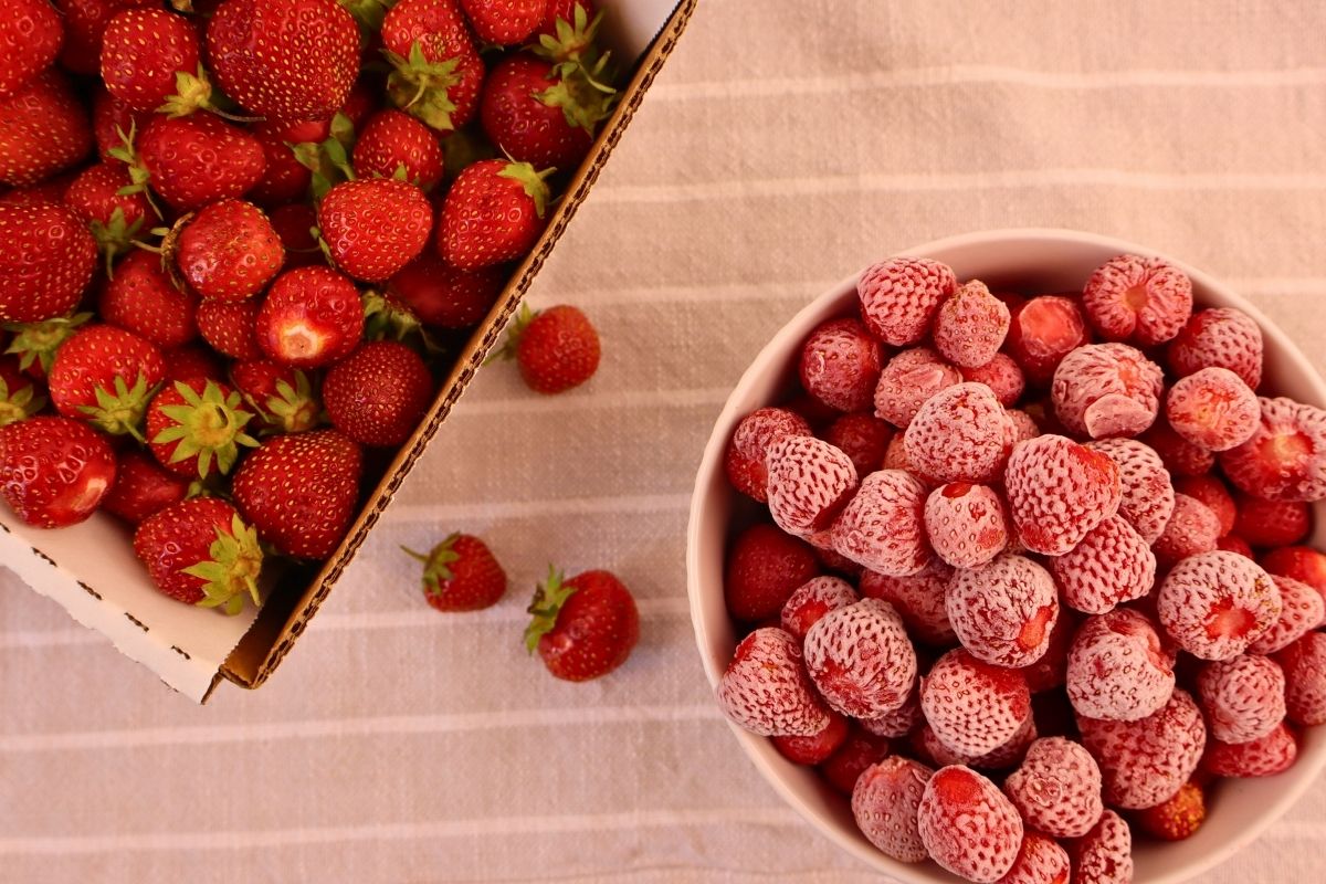Overhead view of a bowl of frozen strawberries beside a box of fresh picked strawberries