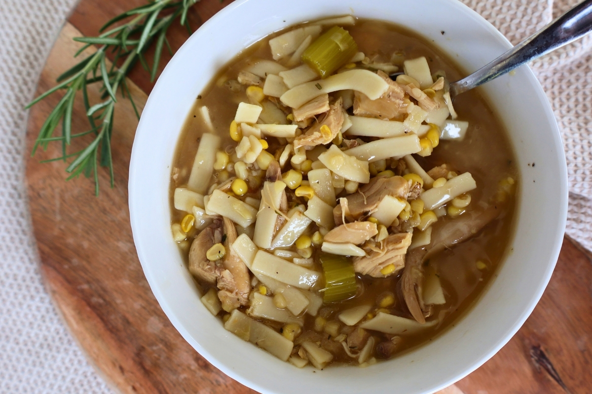 Bowl of homemade gluten free chicken corn soup with a spoon and a fresh rosemary sprig on a wooden board